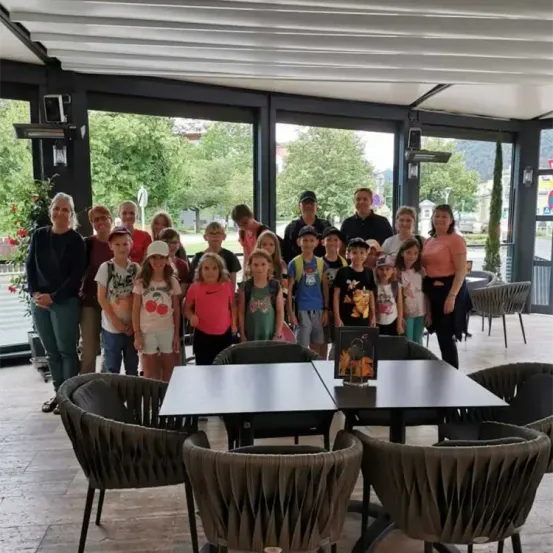 Eine Gruppe von Erwachsenen und Kindern, die zusammen in einem Restaurant stehen. Sie lächeln und posieren für ein Foto. Vor ihnen stehen Stühle und Tische. Das Restaurant hat Glasfenster mit Blick nach draußen.