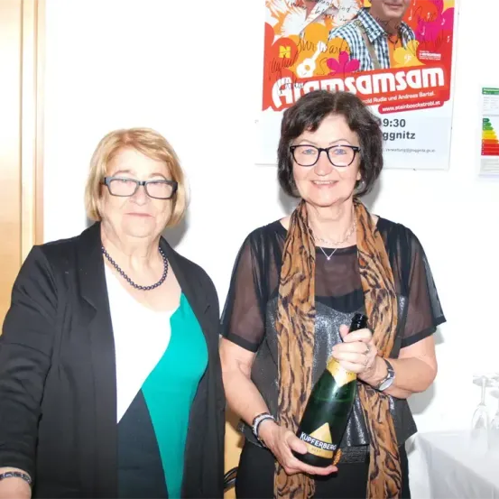 Zwei Frauen stehen zusammen und posieren für ein Foto. Eine von ihnen hält eine Flasche Champagner. Hinter ihnen befindet sich ein Poster an der Wand.