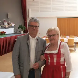 Ein Mann und eine Frau stehen eng beieinander in einer Halle und lächeln. Der Mann trägt eine Brille und einen grauen Anzug, während die Frau ein rotes Kleid trägt. Hinter ihnen sind Tische für eine Veranstaltung mit Stühlen und Blumenarrangements gedeckt.