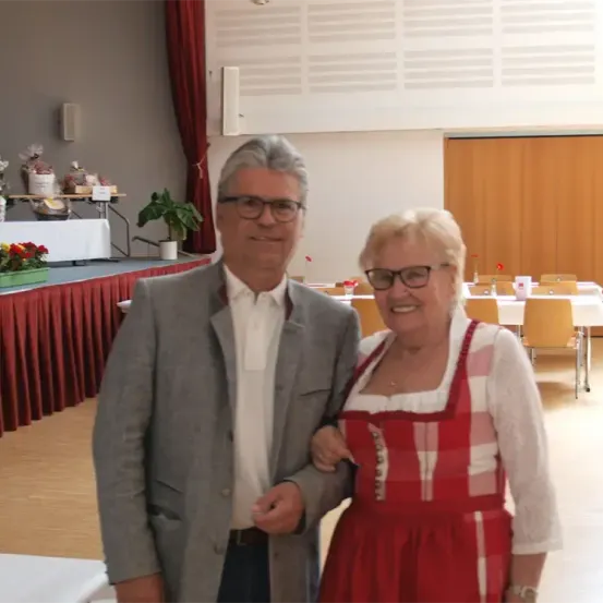 Ein Mann und eine Frau stehen eng beieinander in einer Halle und lächeln. Der Mann trägt eine Brille und einen grauen Anzug, während die Frau ein rotes Kleid trägt. Hinter ihnen sind Tische für eine Veranstaltung mit Stühlen und Blumenarrangements gedeckt.