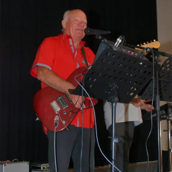 Ein älterer Mann in einem roten Shirt spielt eine rote Gitarre bei einem Konzert, steht vor einem Mikrofon auf einem Ständer.