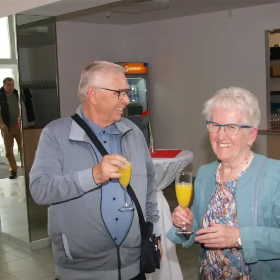 Ein älteres Paar lächelt und hält Gläser mit Orangensaft, steht in einem Raum mit einem Tisch und einem Verkaufsautomaten. Ein Mann wird in einer Glastür im Hintergrund reflektiert.