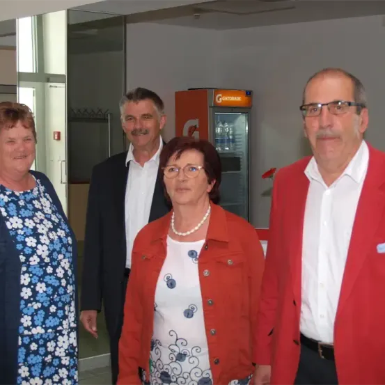 Vier Personen stehen in einem Raum und lächeln für ein Foto. Eine Frau in einem Blumenkleid und einer Perlenkette, ein Mann in einem schwarzen Anzug, eine Frau in einer roten Jacke und ein Mann in einem roten Blazer.