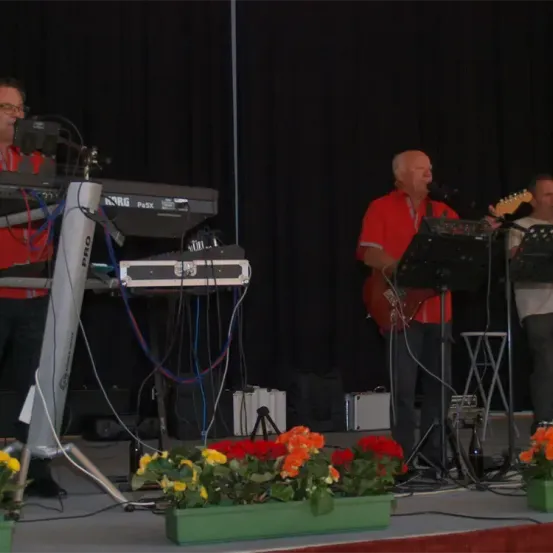 Eine Band spielt auf der Bühne mit schwarzem Vorhang im Hintergrund. Ein Mann in einem roten Hemd spielt Gitarre und singt, während ein anderer Mann das Keyboard spielt. Es gibt Topfblumen auf der Bühne.
