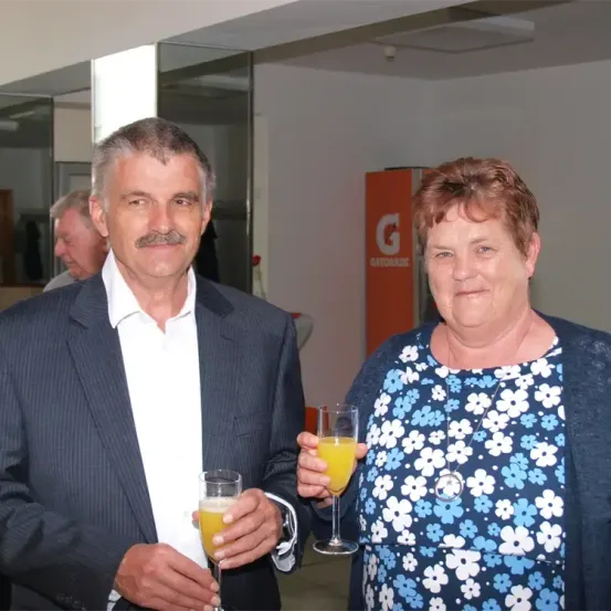 Ein älterer Mann und eine ältere Frau halten Orangengetränke und lächeln für ein Foto. Der Mann hat einen Schnurrbart und die Frau eine Halskette. Hinter ihnen steht ein Mann in der Nähe einer Glaswand.