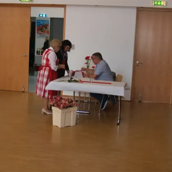 Ein Mann und zwei Frauen stehen an einem Tisch mit Blumen und Dokumenten. Die Frau in Rot und Weiß hält ein Dokument, während der Mann und die andere Frau es betrachten.