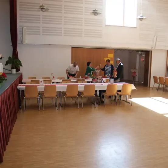 Mehrere Menschen versammeln sich um einen langen Tisch in einem Raum. Einige stehen, andere sitzen. Der Tisch ist mit einem weißen Tuch bedeckt. Es gibt Stühle auf beiden Seiten.