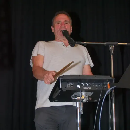 Ein Mann steht auf der Bühne, hält einen Drumstick und ein Mikrofon in der Hand, wahrscheinlich beim Auftritt oder Üben. Er trägt ein weißes T-Shirt und graue Hose.