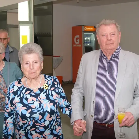 Ein älteres Paar steht Hand in Hand. Die Frau trägt eine Bluse mit Blumenmuster und eine Perlenkette. Der Mann hat ein gestreiftes Hemd und hält ein Glas Orangensaft. Hinter ihnen steht eine Maschine mit der Aufschrift Gatorade.