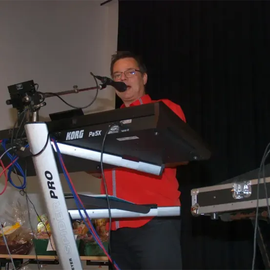 Ein Mann mit Brille und rotem Shirt steht vor einem Korg Pa5X Keyboard. Er singt in ein Mikrofon, während er das Keyboard hält.