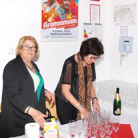 Zwei Frauen stehen hinter einem Tisch voller Gläser und einer Champagnerflasche. Sie sind vor einem Poster mit einem Datum und einem Männergesicht. Die Frau auf der linken Seite hält eine Handtasche. Die Frau auf der rechten Seite trägt eine Brille und einen Schal.