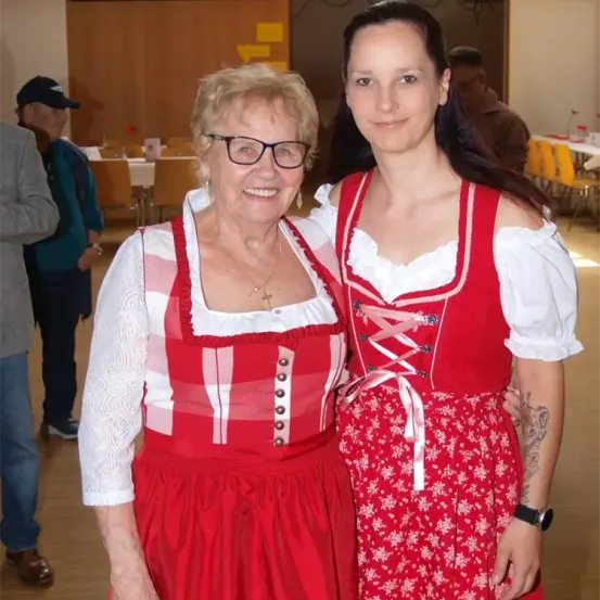 Zwei Frauen in traditioneller deutscher Kleidung, eine ältere und eine jüngere, posieren für ein Foto. Der Hintergrund zeigt einen Raum mit Stühlen und Tischen.
