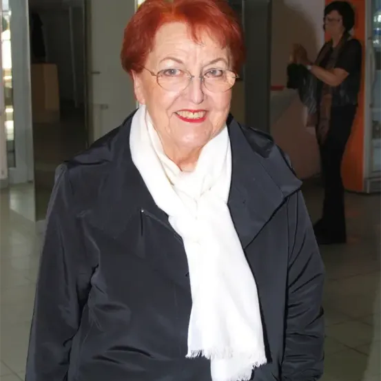 Eine ältere Frau mit roten Haaren, Brille und weißem Schal lächelt in einem Innenkorridor. Eine Person steht hinter ihr und schaut nach unten.