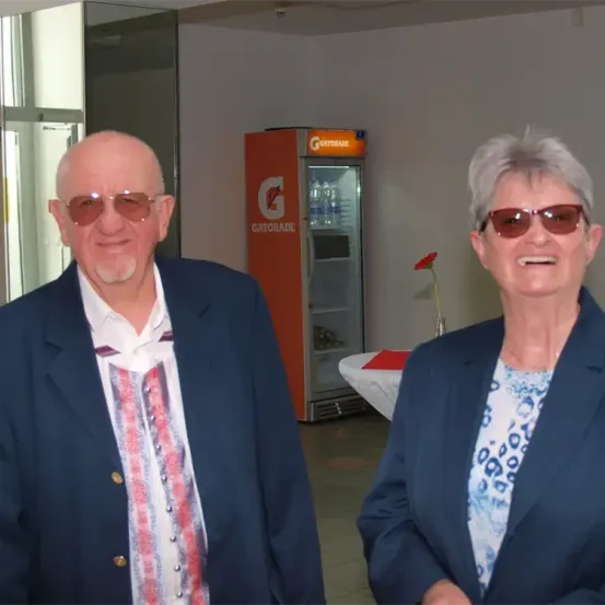 Zwei Menschen lächeln für die Kamera. Der Mann hat einen Bart und trägt eine Sonnenbrille und einen Anzug. Die Frau trägt eine Sonnenbrille, einen Blazer und eine Bluse mit Blumenmuster. Dahinter steht ein Gatorade-Kühlschrank.