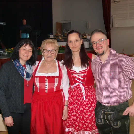 Vier Personen in traditioneller bayerischer Kleidung posieren für ein Foto. Zwei Frauen tragen rote Kleider, während der Mann rechts ein kariertes Hemd und grüne Hosen trägt. Hinter ihnen ist eine Bühneneinrichtung zu sehen.