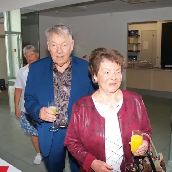 Ein älteres Paar steht in einer Lobby, hält Weingläser mit Orangensaft und lächelt in die Kamera, während eine Frau in einem weißen Shirt hinter ihnen geht.