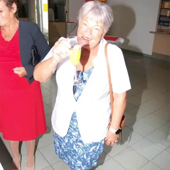 Zwei Frauen stehen in einem Raum; eine trinkt aus einem Weinglas. Die Frau auf der linken Seite trägt ein rotes Kleid und eine Halskette. Die Frau auf der rechten Seite trägt ein weißes Hemd und trägt eine braune Handtasche.