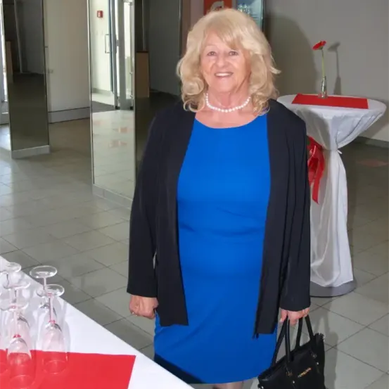 Eine Frau in einem blauen Kleid und einer schwarzen Jacke steht vor einem Tisch mit einer Vase und einem roten Band. Sie hält eine schwarze Handtasche.