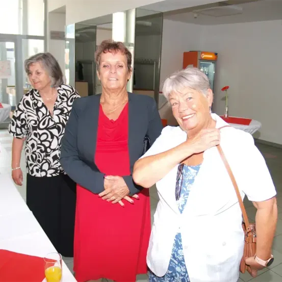 Drei Frauen stehen zusammen, zwei lächeln in die Kamera. Die mittlere Frau trägt ein rotes Kleid, während die anderen Schwarz und Weiß und Weiß und Blau tragen. Dahinter steht ein Tisch mit einem roten Tuch und einer Blume.