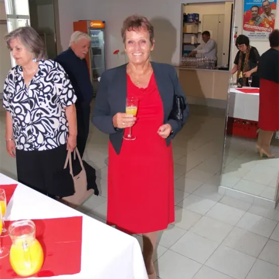 Eine Indoor-Veranstaltung mit mehreren Personen, eine Frau in einem roten Kleid hält ein Glas Orangensaft, andere stehen oder sitzen herum.