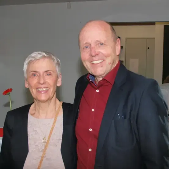 Ein Mann und eine Frau lächeln und stehen einander nahe, wahrscheinlich bei einer Veranstaltung. Der Mann trägt ein rotes Hemd, während die Frau eine Halskette und einen Blazer trägt. Hinter ihnen befindet sich eine Wand mit einer roten Blume auf der linken Seite.