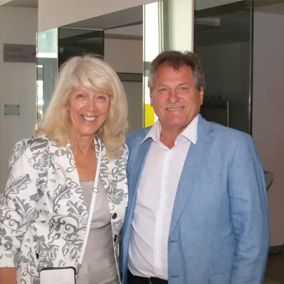 Ein Mann und eine Frau stehen nah beieinander, lächelnd, in einem modernen Innenraum. Die Frau trägt einen gemusterten Blazer und der Mann einen hellblauen Anzug. Hinter ihnen befindet sich eine Glaswand und ein Schild, das 'Zugang Toiletten' anzeigt.