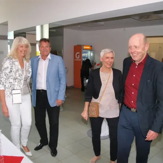 Vier Personen, zwei Männer und zwei Frauen, stehen in einem Raum. Eine Frau trägt eine braune Handtasche. Der Raum hat weiße Fliesen und einen roten Kühlschrank im Hintergrund.