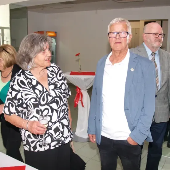 Drei ältere Erwachsene stehen in einem Raum zusammen. Die Frau hält ein Weinglas. Der Mann neben ihr trägt eine Brille und einen blauen Blazer. Ein weiterer Mann hinter ihnen trägt eine Krawatte. Ein Tisch mit einer Blume und einem roten Band steht hinter ihnen.