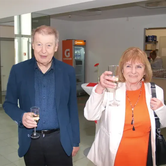 Ein älterer Mann und eine ältere Frau stehen in einem Flur, lächeln und halten Weingläser. Der Mann trägt einen blauen Anzug und die Frau trägt einen weißen Mantel und ein orangefarbenes Kleid. Hinter ihnen sind ein Kühlschrank und ein Tisch mit einem Blumenvase zu sehen.