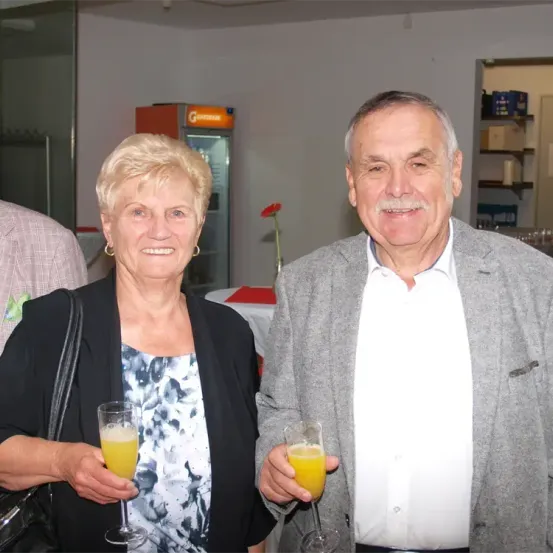 Ein Mann und eine Frau stehen nebeneinander und halten Gläser mit Orangensaft. Sie lächeln und scheinen sich bei einer sozialen Veranstaltung zu befinden. Dahinter steht ein Kühlschrank mit einer Blume und einem Regal mit Lagerboxen.