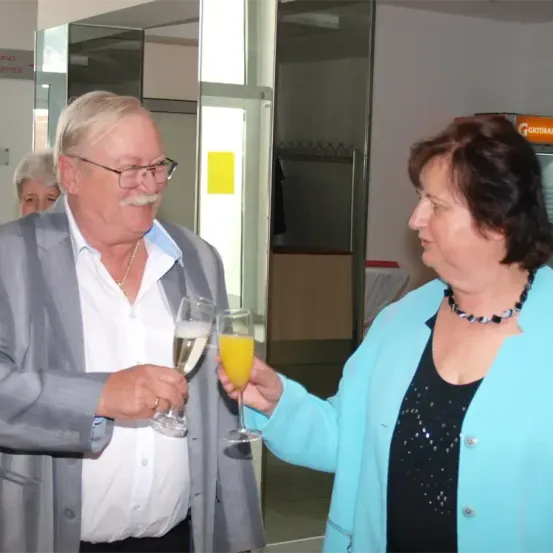 Ein älterer Mann und eine ältere Frau halten Weingläser mit Getränken darin. Der Mann trägt einen Anzug und eine Brille, und die Frau trägt eine blaue Jacke. Sie sind in einem Raum mit Glaswänden.