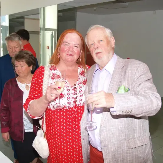 Ein älterer Mann und eine ältere Frau lächeln und halten Weinglas in der Hand in einem Raum mit anderen Leuten im Hintergrund.