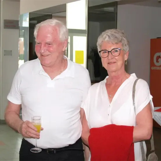 Ein älterer Mann und eine ältere Frau stehen drinnen zusammen. Der Mann hält ein Glas Orangensaft und lächelt. Die Frau trägt eine Brille und eine Halskette und hält ein rotes Tuch. Hinter ihnen befindet sich ein gelbes Schild und eine Glaswand.