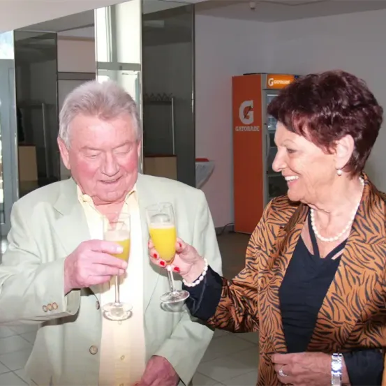 Ein älterer Mann und eine ältere Frau halten Gläser mit Orangensaft, lächeln und stehen in einem Raum. Der Mann trägt einen Anzug und die Frau ein Tigerfell-Blazer. Ein Gatorade-Automat steht im Hintergrund.