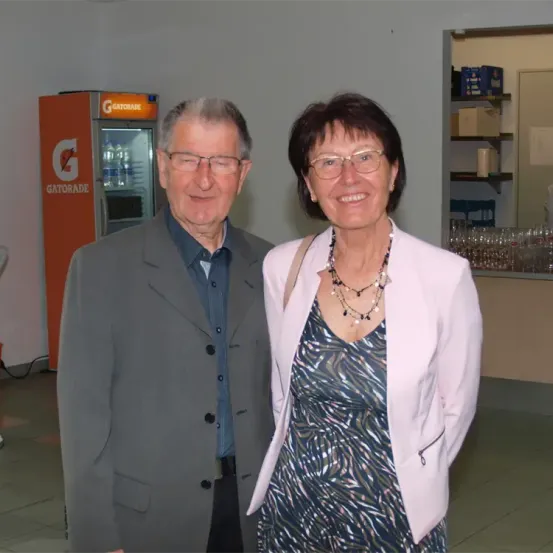 Ein älterer Mann und eine ältere Frau stehen lächelnd zusammen. Der Mann trägt einen grauen Anzug, und die Frau trägt eine rosa Jacke über einem gemusterten Kleid. Hinter ihnen steht ein Gatorade-Automat.