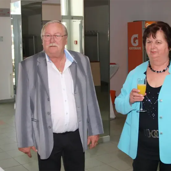 Ein älterer Mann und eine ältere Frau stehen in einem Flur. Der Mann trägt einen grauen Anzug und ein weißes Hemd, und die Frau trägt einen blauen Blazer und ein schwarzes Oberteil. Beide halten Gläser, wobei das der Frau ein gelbes Getränk enthält. Hinter ihnen befindet sich ein Spiegel und ein Schild mit dem Buchstaben 'G'.