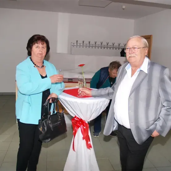 Ein älteres Paar stößt mit Weinglas an einem dekorierten Tisch an. Hinter ihnen beugt sich ein Mann in einer grünen Jacke. Der Raum ist gut beleuchtet und sauber.