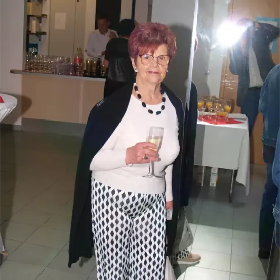 Eine ältere Frau mit rosa Haaren und Brille steht vor einem Spiegel bei einer Veranstaltung und hält ein Glas Wein. Im Hintergrund sehen sich verschwommene Personen, eine Bartheke und ein Tisch mit Getränken.