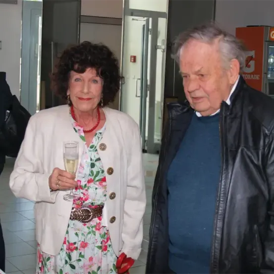 Eine ältere Frau mit einem Weinglas und ein älterer Mann in einer schwarzen Jacke stehen neben ihr in einem Flur.