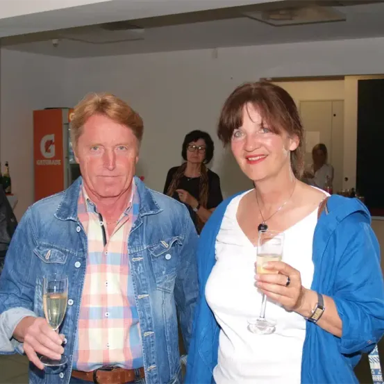 Ein Mann und eine Frau, lächelnd und mit Weinglas in der Hand, stehen eng beieinander in einem Raum. Ein Gatorade-Automat und eine verschwommene Frau sind hinter ihnen.