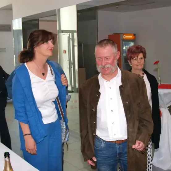 Ein Mann mit Bart und Schnurrbart steht neben einer Frau in einem blauen Kleid. Hinter ihnen steht eine weitere Frau mit Brille in der Nähe eines Tisches mit einer Blume.