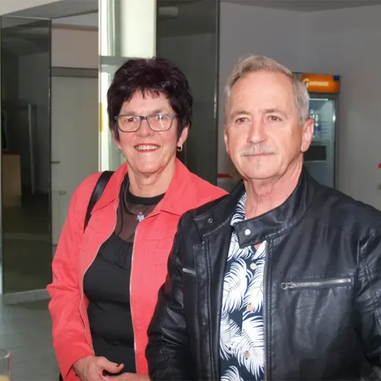 Ein älterer Mann und eine ältere Frau lächeln und stehen eng beieinander in einem Raum. Die Frau trägt eine rote Jacke und eine Brille. Der Mann trägt eine schwarze Lederjacke. Dahinter befinden sich eine Glastür, ein Verkaufsautomat und ein gefliester Boden.