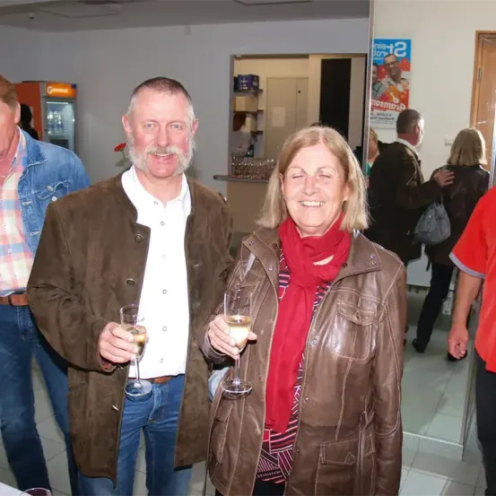 Ein Mann und eine Frau stehen lächelnd nebeneinander und halten Weingläser in einer Hand in einem Raum, in dem andere Leute im Hintergrund stehen.