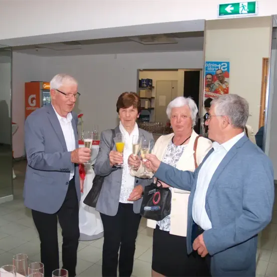 Fünf Personen stehen lächelnd in einem Raum. Vier von ihnen halten Champagnergläser und eine hält eine Handtasche. Der Raum hat weiße Wände und eine weiße Decke. Rechts befindet sich eine Glastür, und ein Plakat ist an der Wand angebracht.