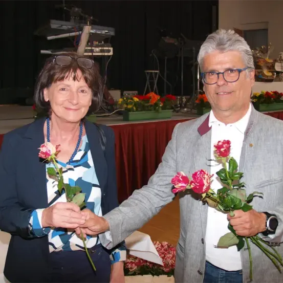 Ein älterer Mann und eine Frau lächeln für ein Foto, während sie Rosen halten. Sie stehen in einem Raum mit einer Bühne und Blumenarrangements im Hintergrund.