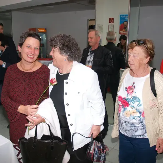 Drei Frauen und zwei Männer stehen in einem Flur, wahrscheinlich bei einer Veranstaltung. Die Frau in der Mitte hält eine Rose und eine weiße Jacke. Hinter ihnen stehen ein Mann in einer schwarzen Jacke und ein anderer Mann in einem schwarzen Hemd.