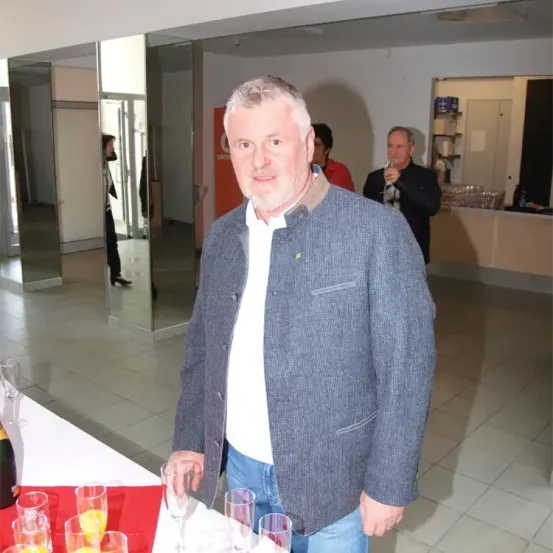 Ein Mann steht vor einem Tisch mit Weingläsern und einer Flasche und lächelt. Hinter ihm stehen andere in einem Raum mit Spiegeln, einer Theke und einem Regal.