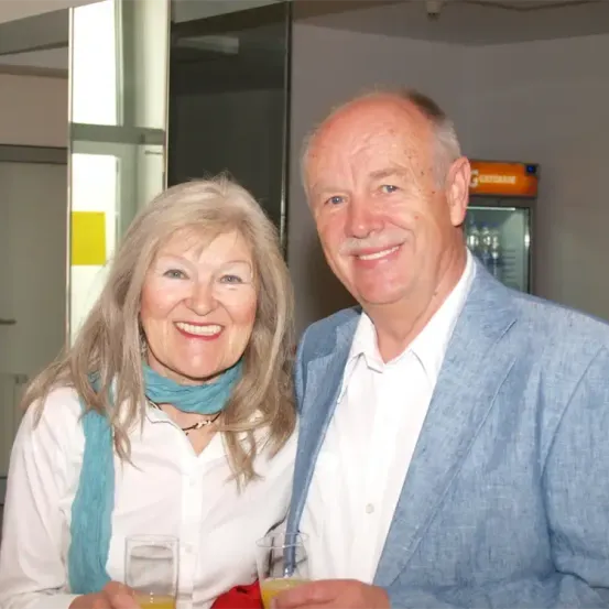 Ein Mann und eine Frau lächeln in die Kamera und halten Gläser mit Orangensaft. Sie stehen eng beieinander. Im Hintergrund ist ein verschwommener Hintergrund mit einem gelben Aufkleber und einem Kühlschrank.