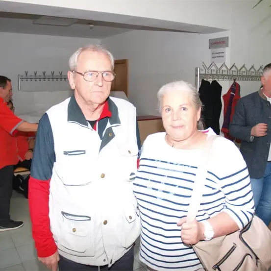 Ein älterer Mann und eine ältere Frau stehen in einem Raum zusammen, mit einem Mann in einem roten Shirt und einem anderen Mann in einem grauen Jackett im Hintergrund.