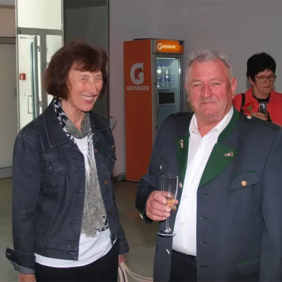 Ein Mann und eine Frau stehen lächelnd in einem Veranstaltungssaal. Der Mann hält ein Weinglas. Hinter ihnen steht ein Verkaufsautomat und eine weitere Frau.